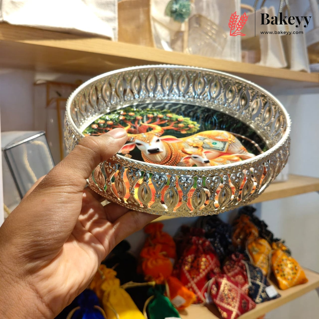 A hand holds a round silver-toned metal tray with ornate cutout patterns and a colorful printed illustration of a deity on the bottom. The background shows wooden shelves with various items in a shop setting, and a 'Bakeyy' watermark is visible in the top right corner.