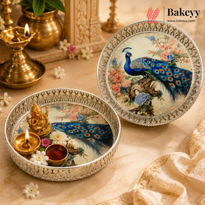 Two ornate round decorative plates with silver borders and peacock illustrations, styled on a beige surface with brass lamps, flowers, and a Bakeyy brand mark in the top right.