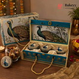 A decorative gift set featuring a large blue and cream peacock-themed box with three matching round tins inside, shown open on a wooden table. A smaller matching box sits behind it, and a small branded Bakeyy logo appears in the top right corner.