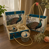 A decorative gift set featuring a blue and cream peacock-themed box with rope handles, an open matching box with a round tin and small jar inside, and a matching tote bag on a wooden table. The set includes printed peacock artwork, cream fabric lining, and glass containers.