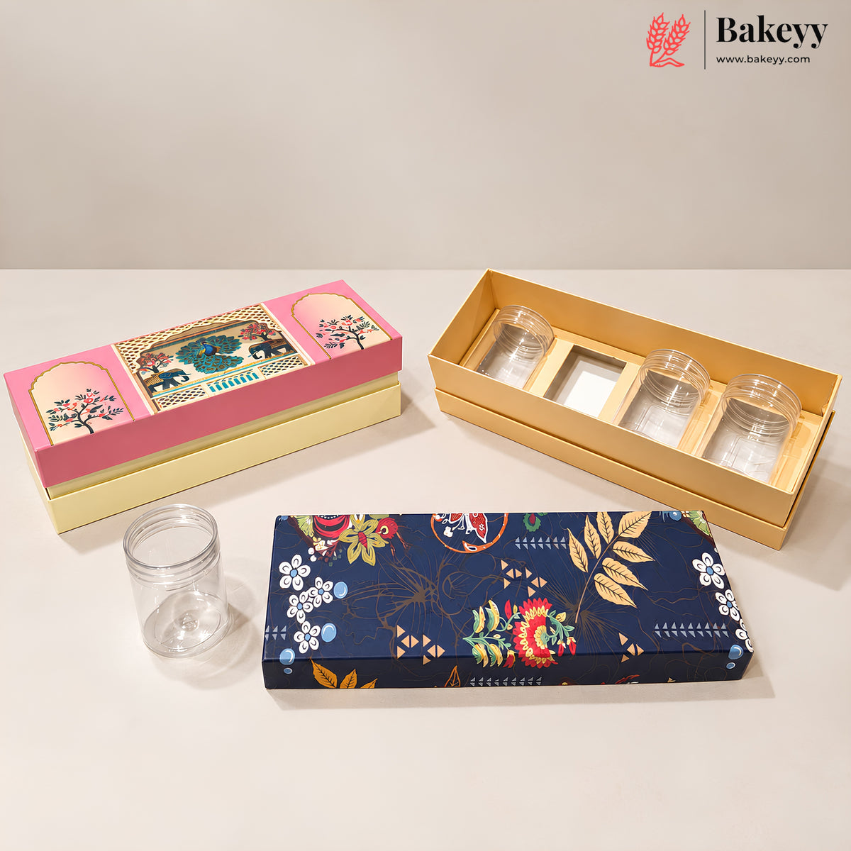Three decorative glass jars with lids displayed on a light beige surface against a neutral background. The set includes a pink and cream box with an elephant illustration, an open natural wood box containing three clear glass jars, and a navy blue floral-patterned box with a single clear glass jar in front.