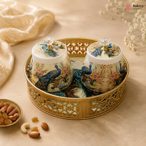 Two decorative ceramic jars with matching lids, decorated with peacock and floral motifs, presented on a gold metal tray. The scene is styled with a gold fabric backdrop, scattered almonds, and a small bowl of dried fruits, with the Bakeyy brand logo in the top right corner.