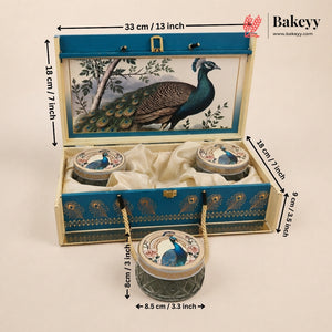 An open decorative Bakeyy gift box for tea or biscuits, shown with dimension callouts. The box is cream-colored wood with a blue lid panel featuring a peacock illustration, velvet lining, and three small round containers attached by rope handles.
