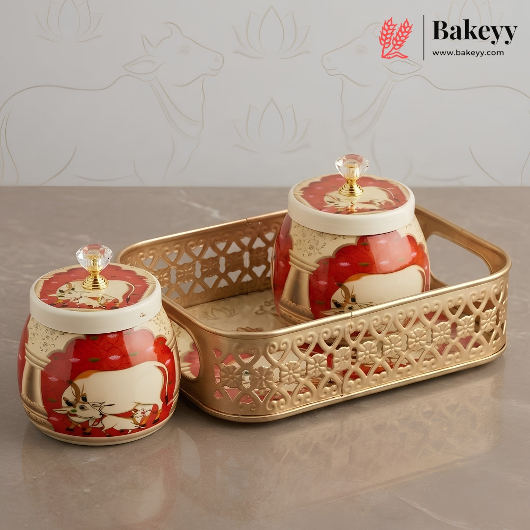 Two decorative ceramic jars with red and cream floral designs and gold lids, displayed on a gold tray against a light gray background with faint cow motifs. The Bakeyy brand logo is visible in the top right corner.