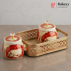 Two decorative ceramic jars with red and cream floral designs and gold lids, displayed on a gold tray against a light gray background with faint cow motifs. The Bakeyy brand logo is visible in the top right corner.