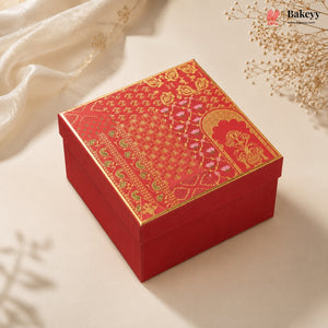 A square red gift box with an ornate gold and pink patterned lid, styled on a light beige surface with soft white fabric and dried floral decorations.