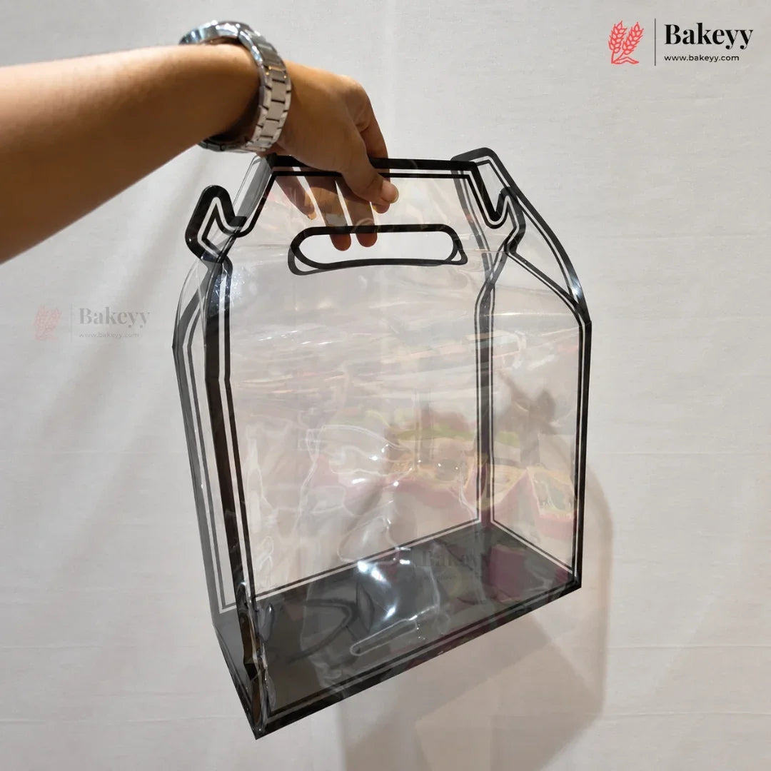 A clear plastic bakery gift bag with a black metal handle frame, held by a person's hand against a white studio background. The Bakeyy logo is visible in the top right corner.