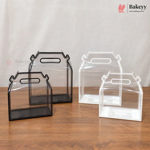 Three clear plastic bakery gift boxes with integrated handles, shown in black and white frames on a wooden table against a neutral fabric backdrop. The Bakeyy logo is visible in the top right corner.