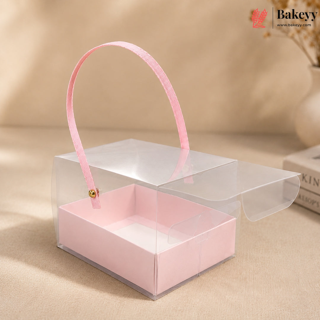 Clear gift box with pink handle and interior on a beige surface, featuring the Bakeyy brand.