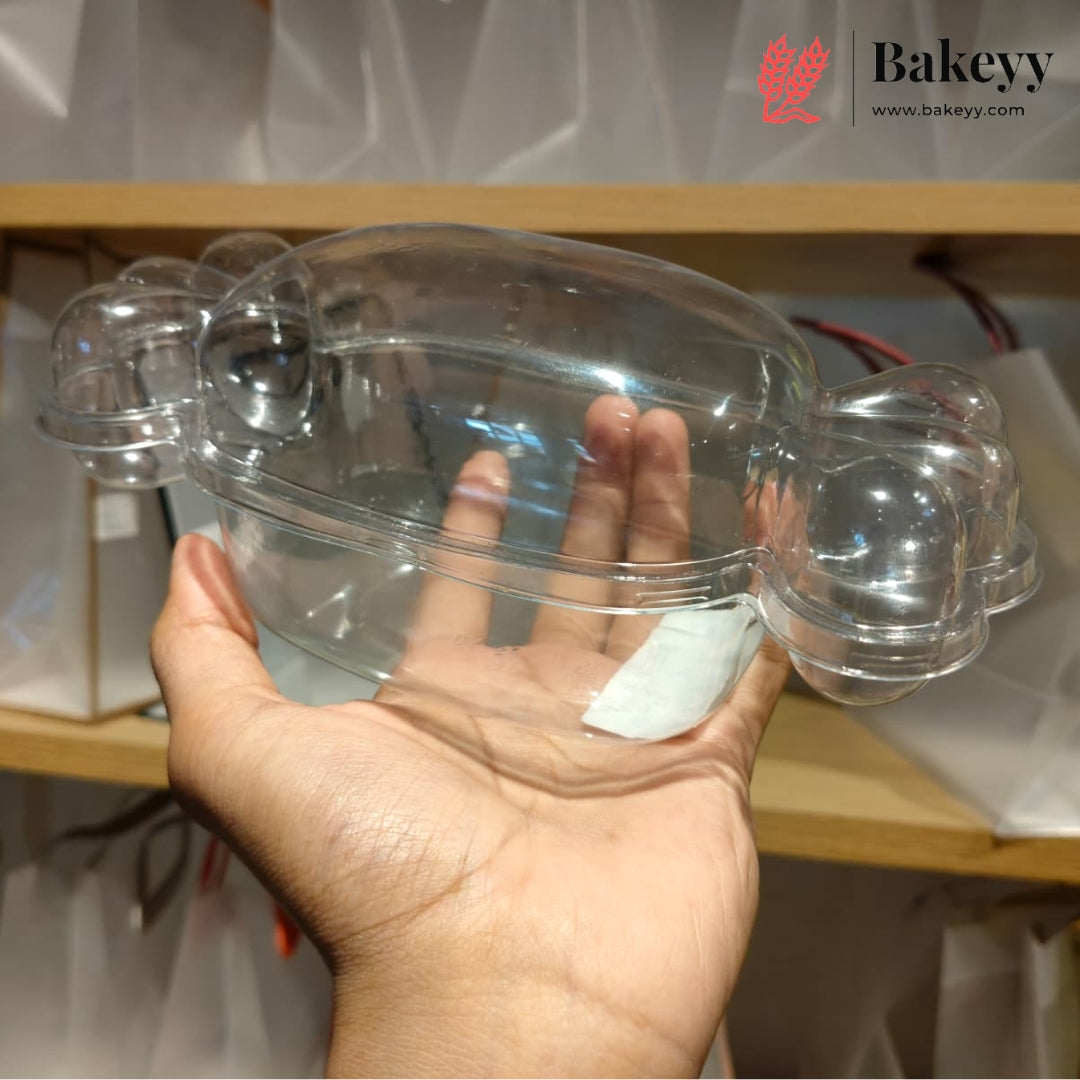 A hand holding a clear plastic bakery mold or tray with two small raised sections on the sides, shown against a shelf background with a Bakeyy logo in the top right.
