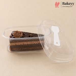 A clear plastic rectangular cake container holding a slice of chocolate cake with whipped cream, shown with its transparent lid leaning against it on a beige background. The Bakeyy logo appears in the top right corner.