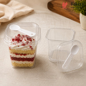 Clear plastic square food containers with white plastic spoons, shown with and without lids on a beige fabric surface. One container is filled with a layered cake dessert, and the brand logo Bakeey appears in the top right corner.