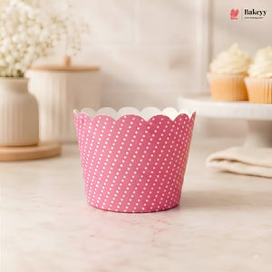 A pink paper cupcake liner with white polka dots and a scalloped edge, shown on a light marble countertop with a blurred bakery background featuring cupcakes and a Bakeyy logo in the top right.