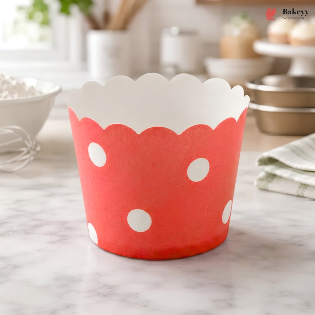 A red paper cupcake liner with white polka dots and a scalloped white inner edge, standing on a marble countertop in a softly blurred kitchen setting.