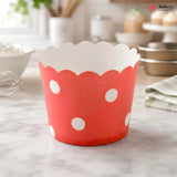 A red paper cupcake liner with white polka dots and a scalloped white inner edge, standing on a marble countertop in a softly blurred kitchen setting.