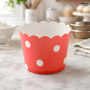 A red paper cupcake liner with white polka dots and a scalloped white inner edge, standing on a marble countertop in a softly blurred kitchen setting.