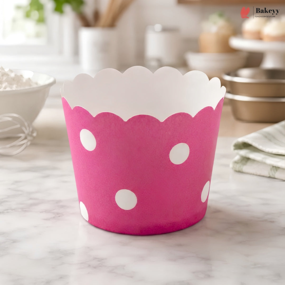 A pink paper cupcake liner with white polka dots and a scalloped white inner edge, standing on a marble countertop in a softly blurred kitchen setting.