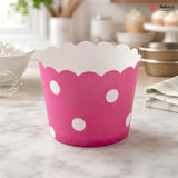 A pink paper cupcake liner with white polka dots and a scalloped white inner edge, standing on a marble countertop in a softly blurred kitchen setting.