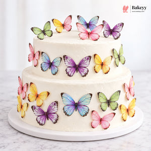 A two-tier white frosted cake decorated with colorful butterfly toppers in pink, purple, blue, yellow, and green, presented on a white cake board against a light background. The BakeDesigns logo is visible in the top right corner.