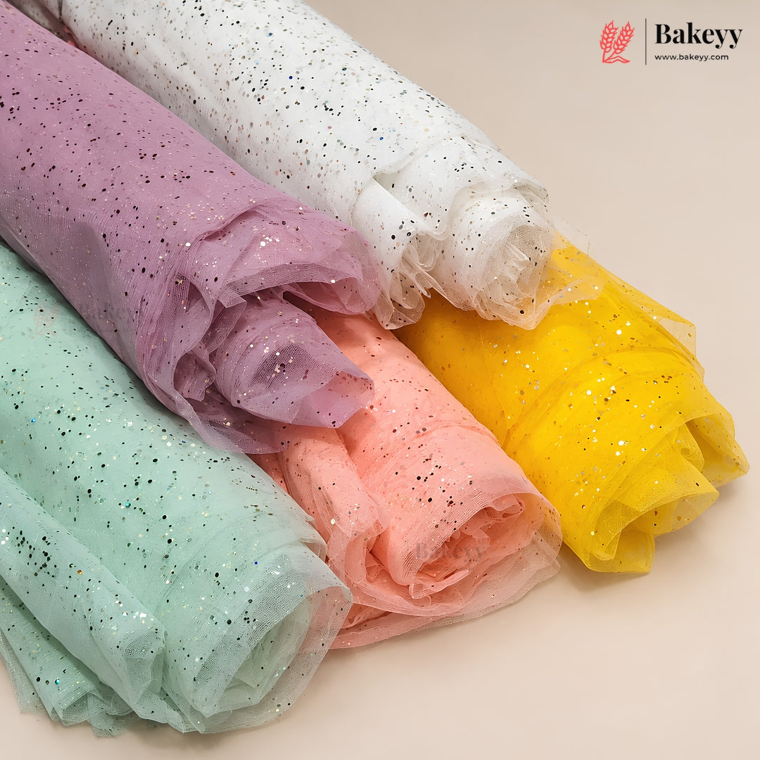 Four rolls of sheer tulle fabric with embedded glitter, shown in pastel shades of purple, white, yellow, and mint green on a light beige background. The Bakeyy logo is visible in the top right corner.