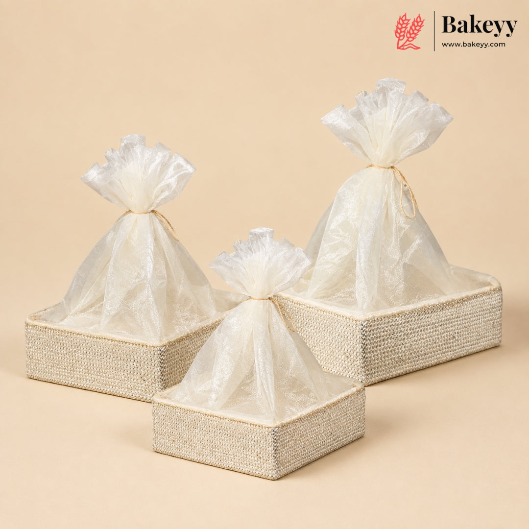 Three woven rectangular gift boxes in beige and cream, each holding a translucent cellophane drawstring bag tied at the top, arranged on a warm beige background. A Bakeyy logo appears in the upper right corner.