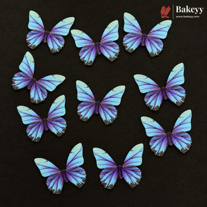 Ten butterfly-shaped cake toppers in shades of purple, blue, and white, arranged on a dark textured background. The Bakeyy brand logo is visible in the top right corner.
