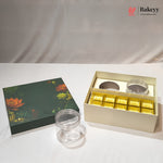 A bakery gift set featuring a dark green lotus-themed box lid, an open cream-colored box containing gold foil-wrapped treats and two clear round jars, and one loose clear jar on a white surface.