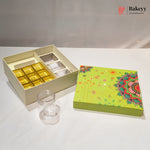 A bakery gift box set on a white surface, featuring an open cream-colored box with gold and white compartments, a clear plastic jar, and a decorative lime green lid with a colorful mandala pattern. The Bakeyy logo is visible in the top right corner.