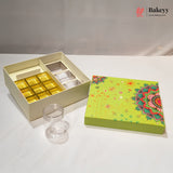 A bakery gift box set on a white surface, featuring an open cream-colored box with gold and white compartments, a clear plastic jar, and a decorative lime green lid with a colorful mandala pattern. The Bakeyy logo is visible in the top right corner.