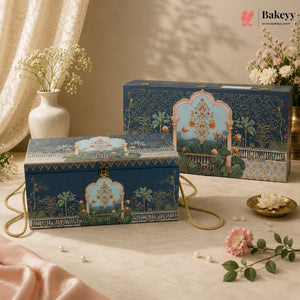 Two decorative blue bakery gift boxes with gold handles and ornate illustrated arch motifs, styled on a beige fabric surface with flowers and draped satin in a warm indoor setting. A Bakeyy watermark/logo appears in the top right.