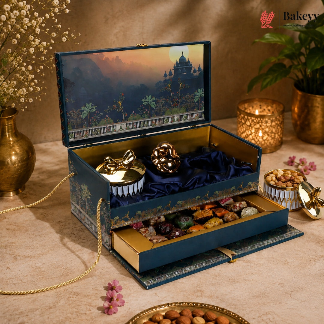 An ornate blue and gold gift box set displayed on a beige tablecloth, featuring a decorative illustrated lid, plush velvet compartments, and an open drawer filled with assorted chocolates and nuts. The scene is styled with candles, flowers, and a plant, and the Bakevy logo appears in the top right corner.