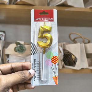 A hand holding a packaged gold number 5 birthday candle with a clear plastic base on a retail display shelf.