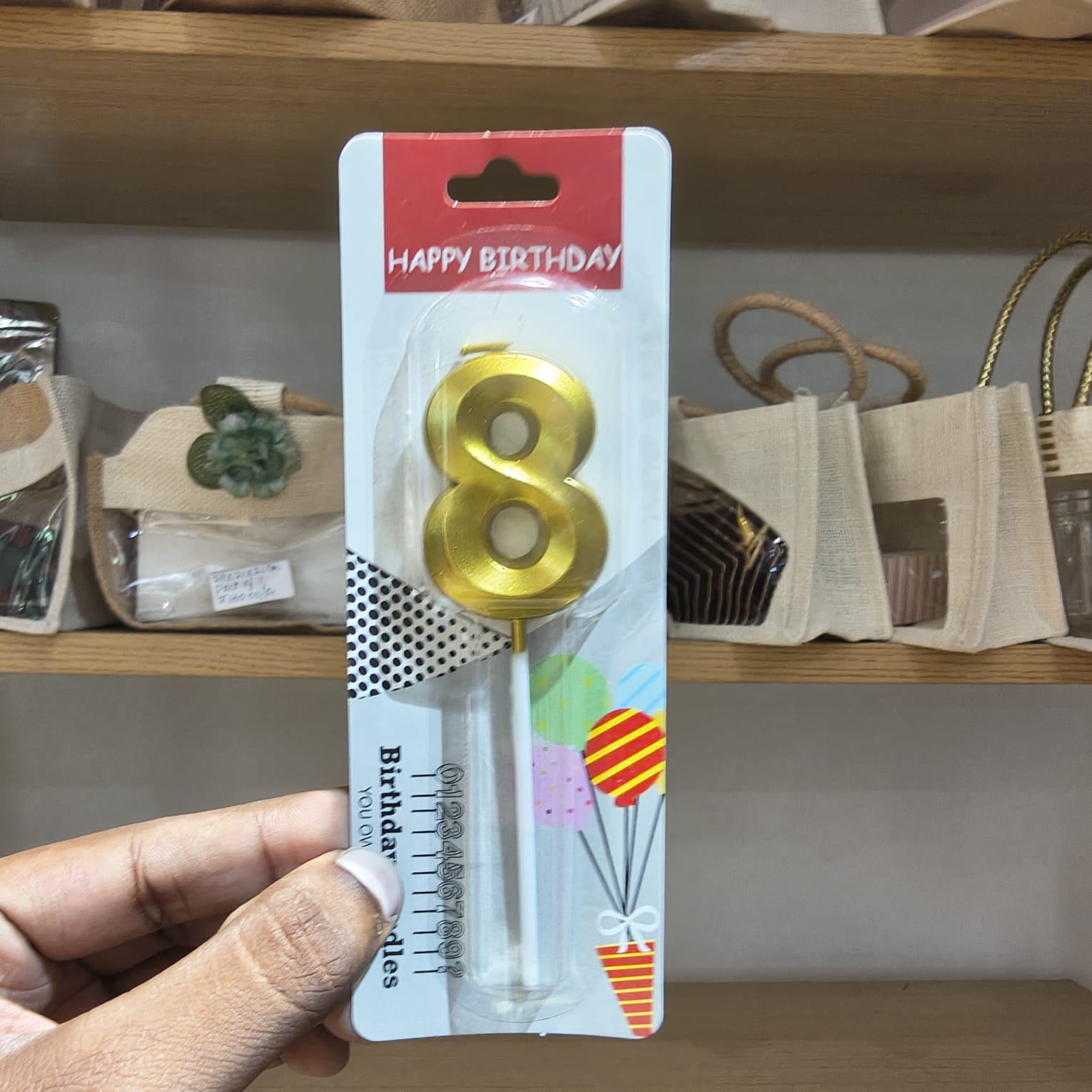 A hand holding a packaged gold number 8 birthday candle on a retail display card, with shelves and bags in the background.