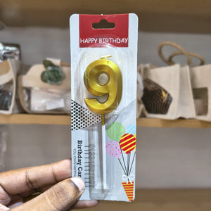 A hand holding a blister pack containing a gold metal number 9 birthday candle, with a red 'HAPPY BIRTHDAY' header and colorful balloon graphics on the card backing.