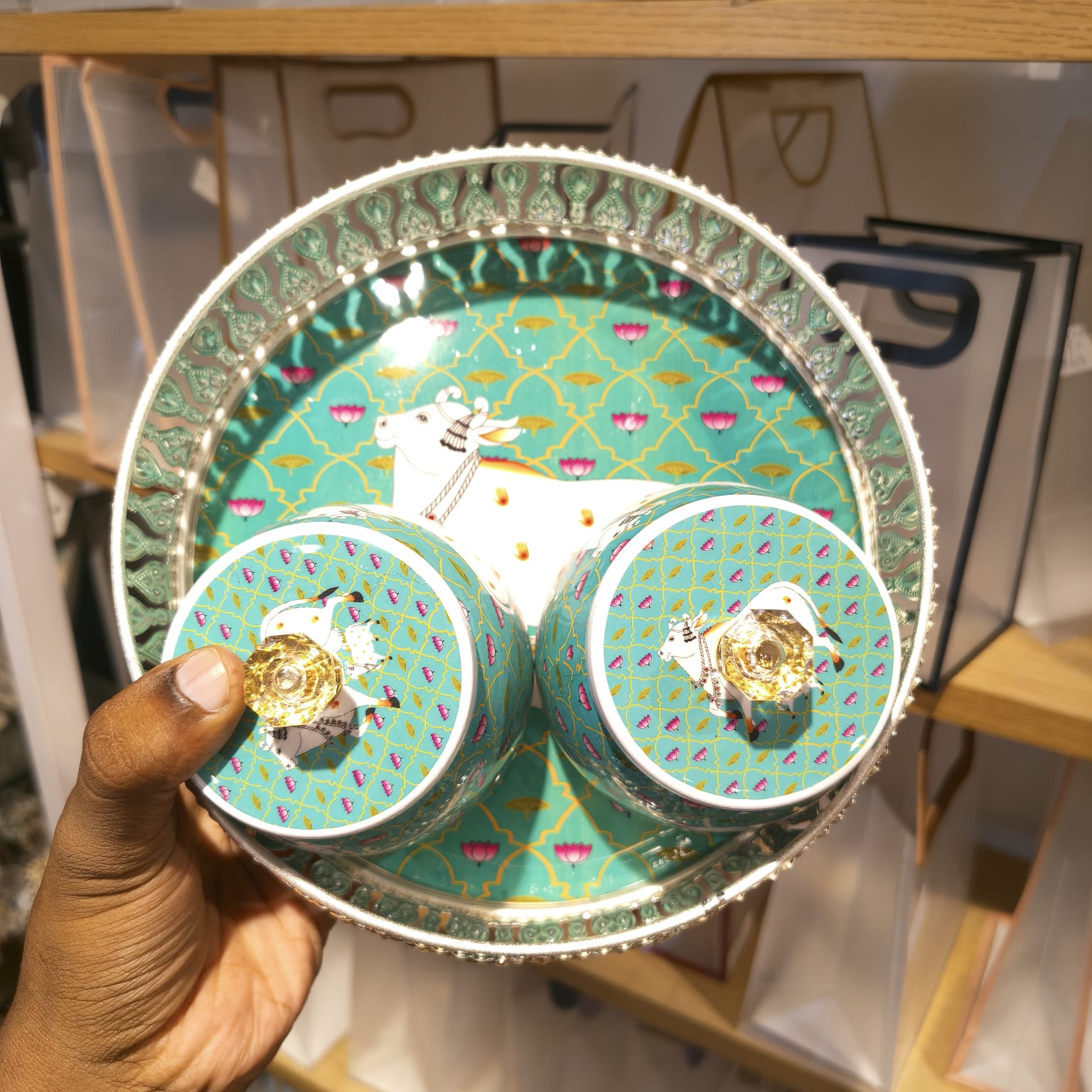 A hand holds a round decorative tray holding two matching small lidded boxes. The items have a turquoise and gold patterned design with small pink and purple accents, photographed in a retail setting with shelves in the background.