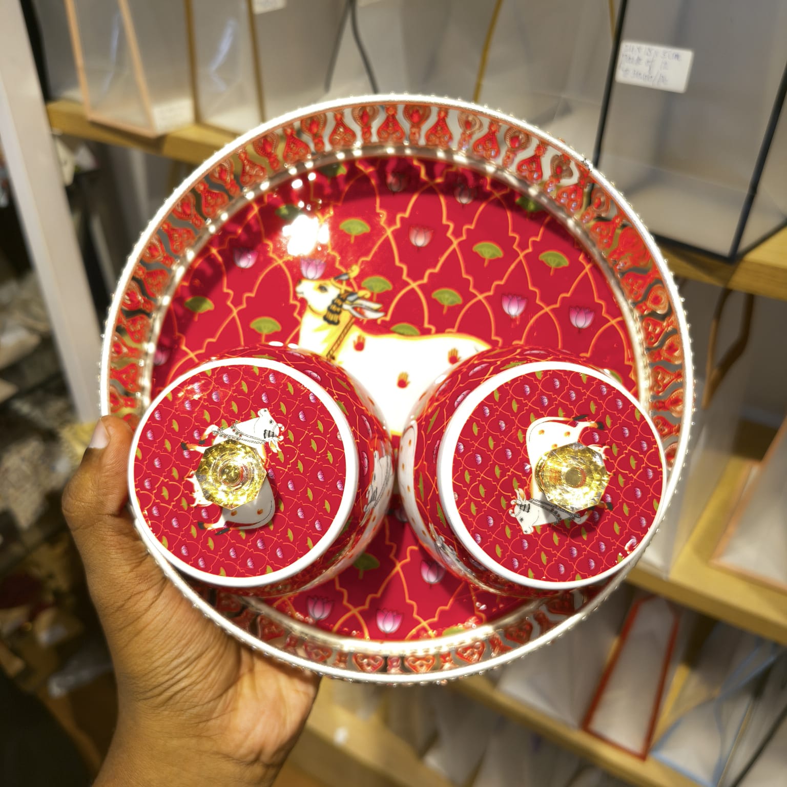 A hand holds a round tray and matching cup set decorated with a festive red and gold pattern, featuring a white stag illustration and small gold clover accents.