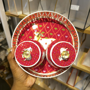 A hand holds a round tray and matching cup set decorated with a festive red and gold pattern, featuring a white stag illustration and small gold clover accents.
