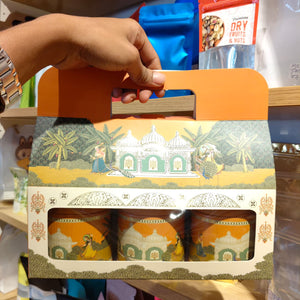 A hand holds an orange-handled gift box containing three decorated ceramic cups with a traditional Indian architectural illustration. The box has a cream and orange printed design with a window showing the cups inside.