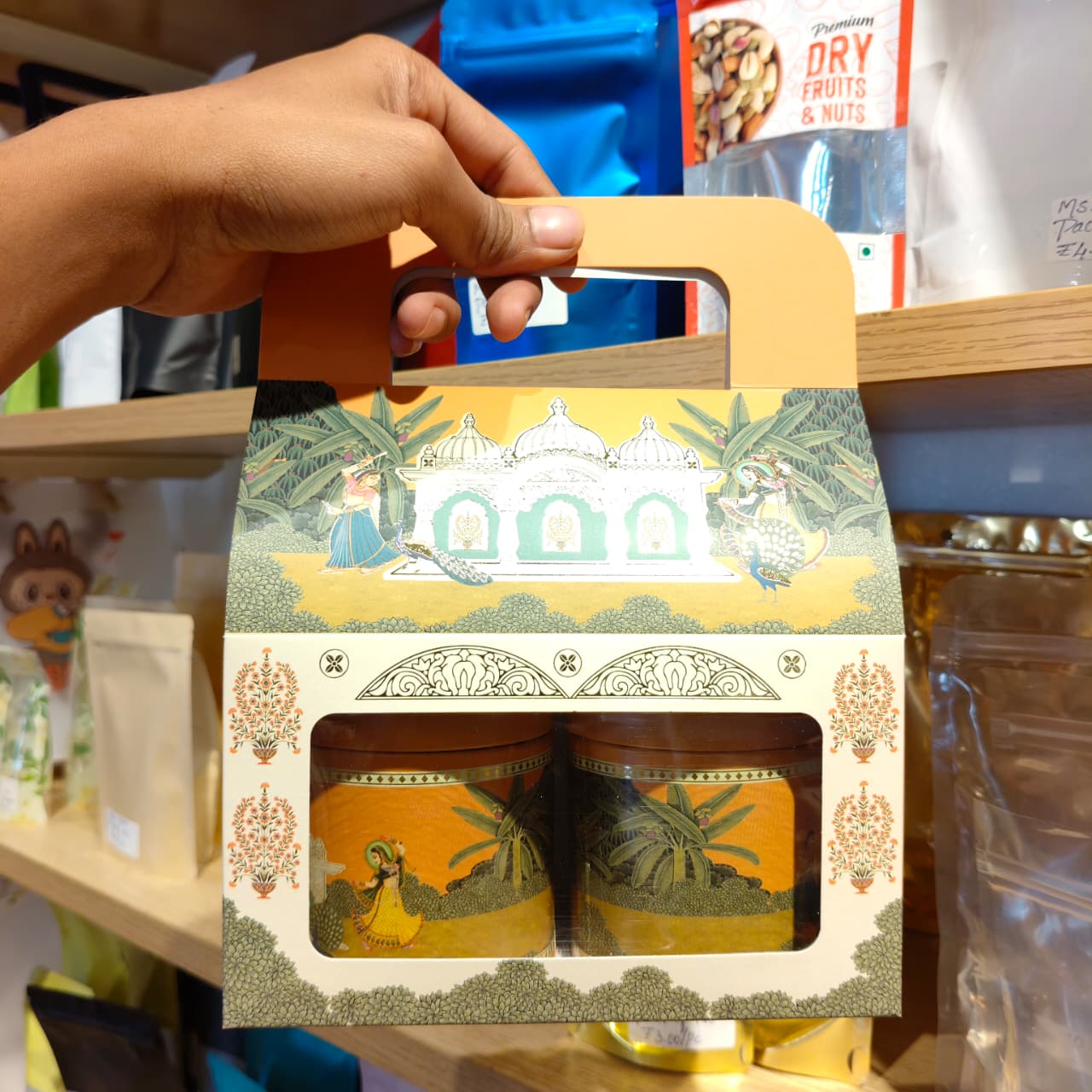 A hand holds a decorative gift box containing two small jars, photographed on a retail shelf. The packaging has a warm yellow and cream color scheme with traditional illustrated motifs and a cutout window showing the jars inside.
