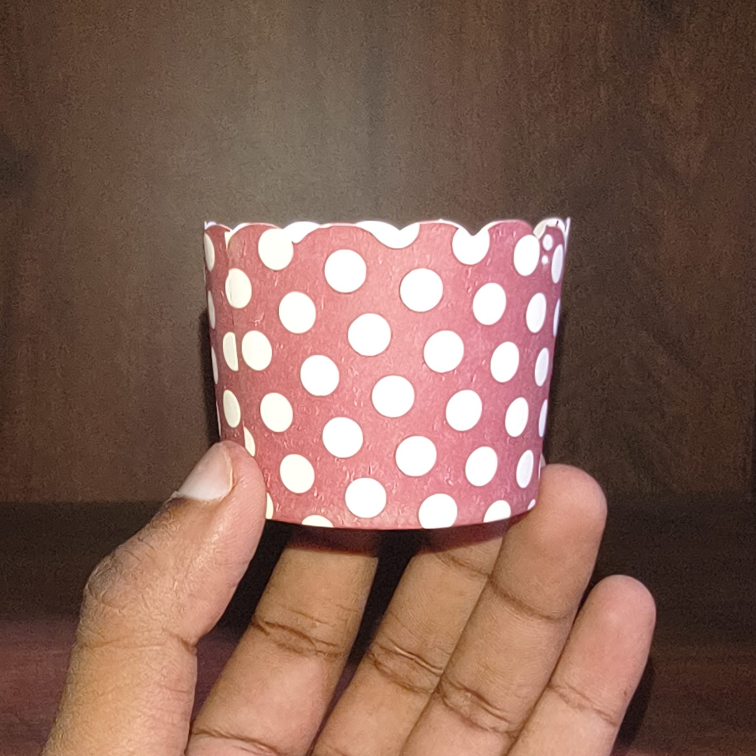 Hand holding a pink paper cup with white polka dots against a brown background