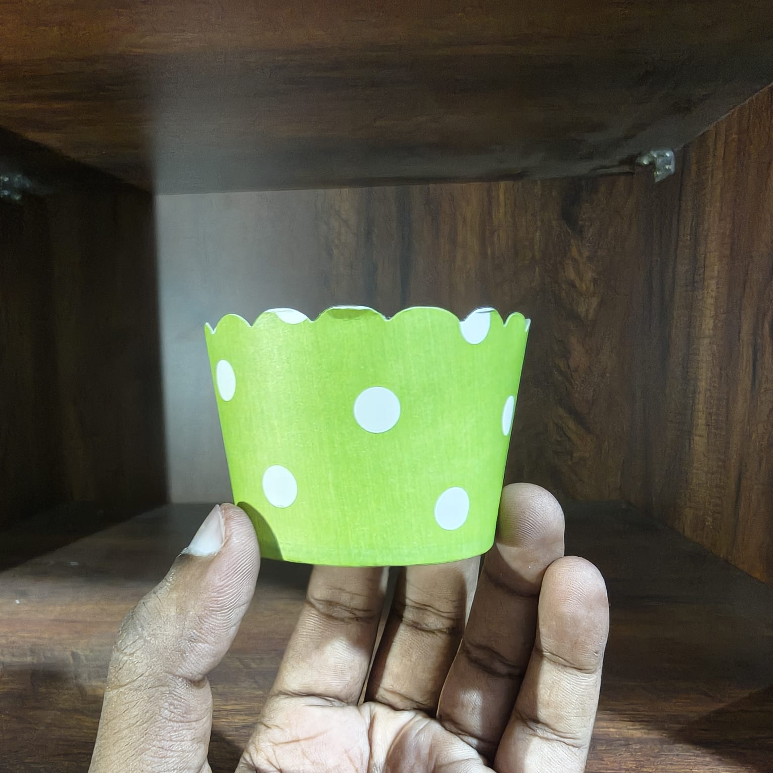 Hand holding a green paper cupcake liner with white polka dots against a wooden background