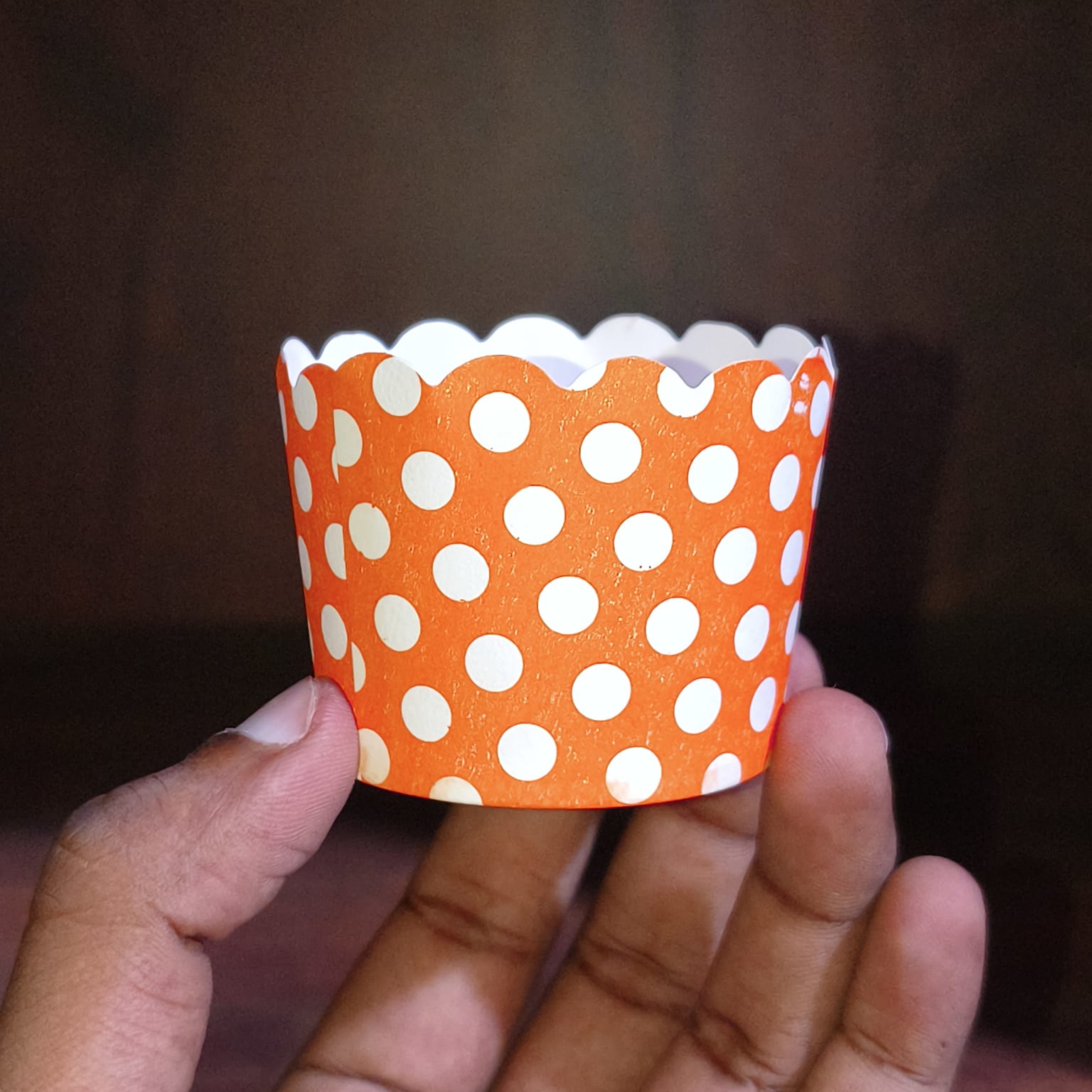 Hand holding an orange paper cup with white polka dots against a dark background
