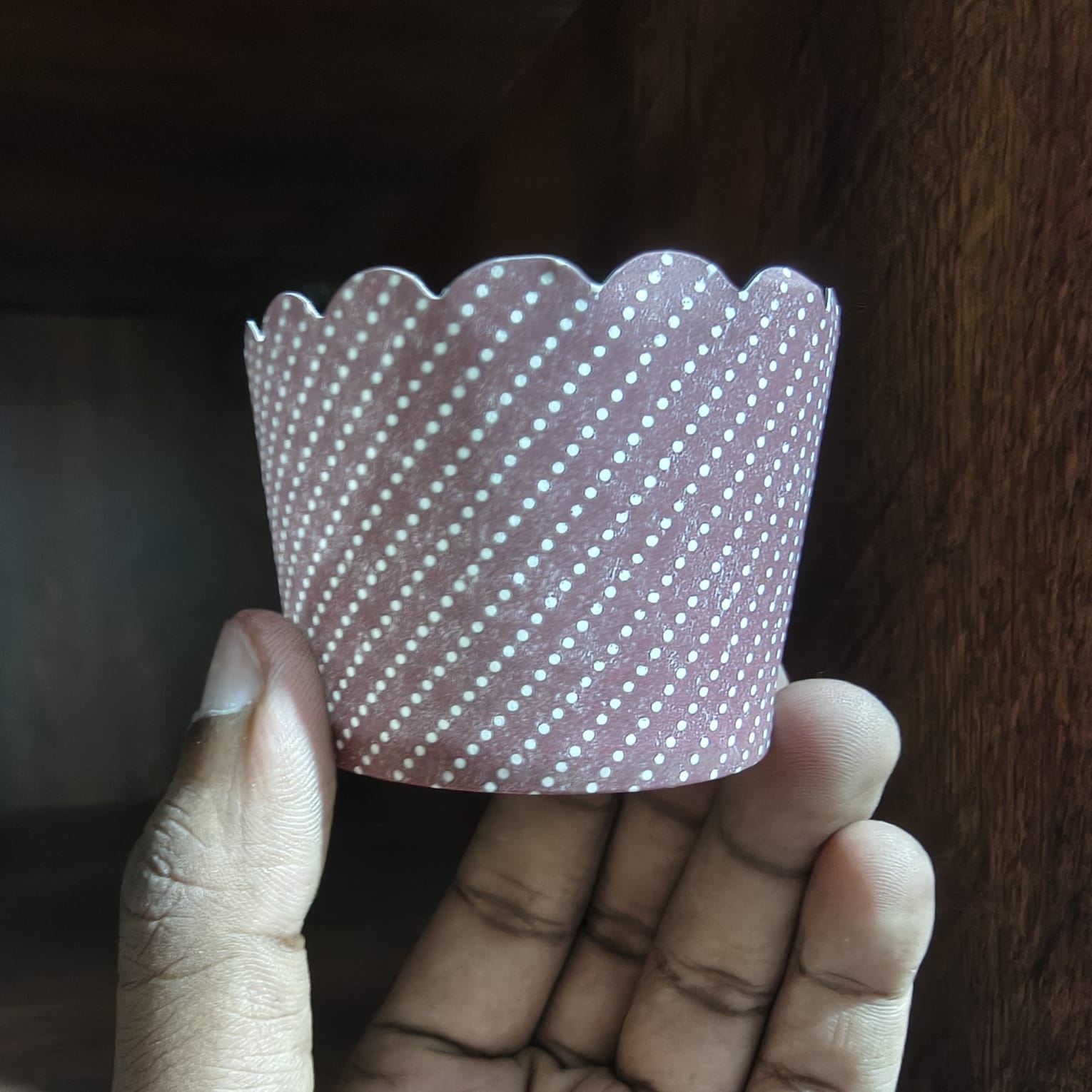 Hand holding a pink cupcake liner with white polka dots against a dark background