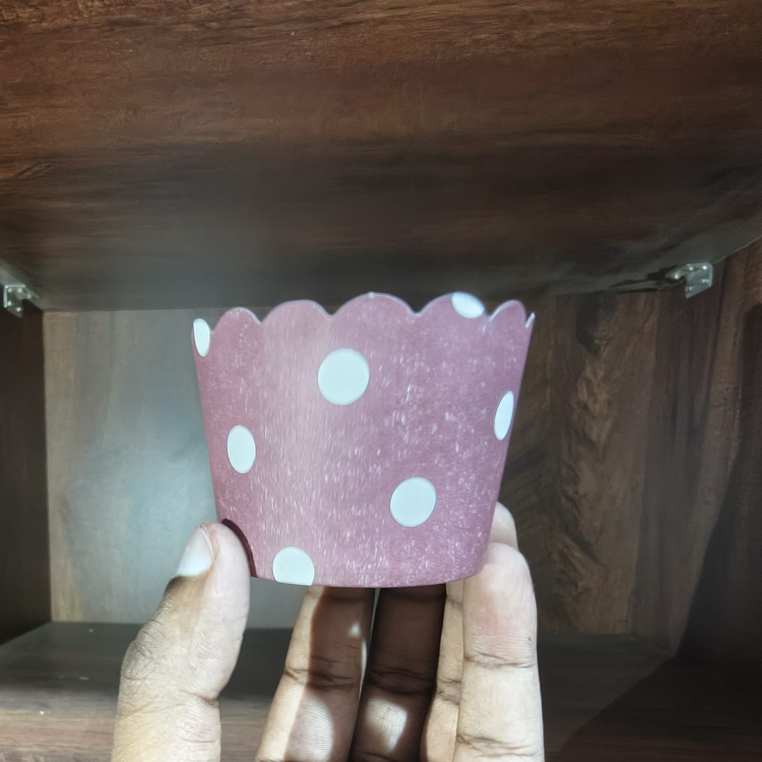 Hand holding a pink paper cupcake liner with white polka dots under a wooden shelf.