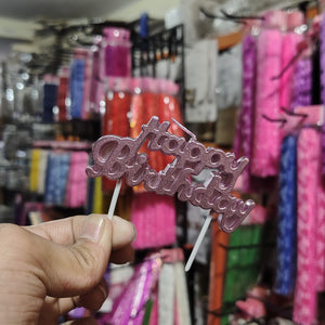 Hand holding a pink 'Happy Birthday' cake topper against a colorful store background.