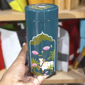 Teal cylindrical container with cow and floral design held by a hand against a blurred background