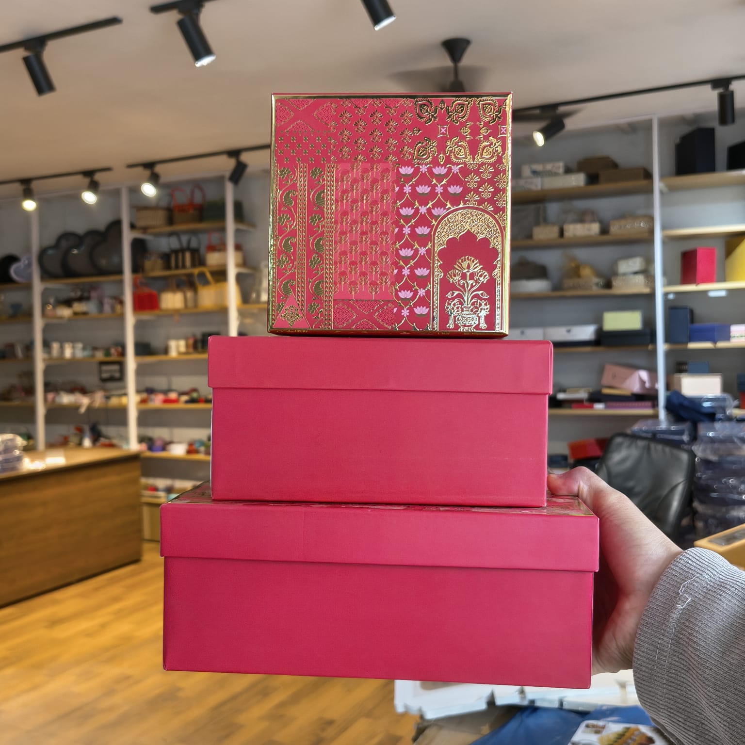Three stacked gift boxes in a retail store setting, with the top box featuring a red and gold traditional patterned design and the bottom two boxes in solid bright pink. A hand is holding the stack on the right side.