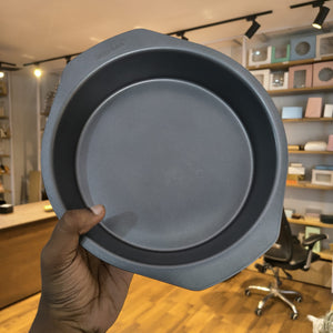 A person's hand holding a round gray non-stick baking pan with a dark interior, set against a blurred background of a retail store interior.