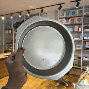 A hand holding a round silver metal baking pan with a raised rim, photographed in a retail store with shelves and fixtures in the background.