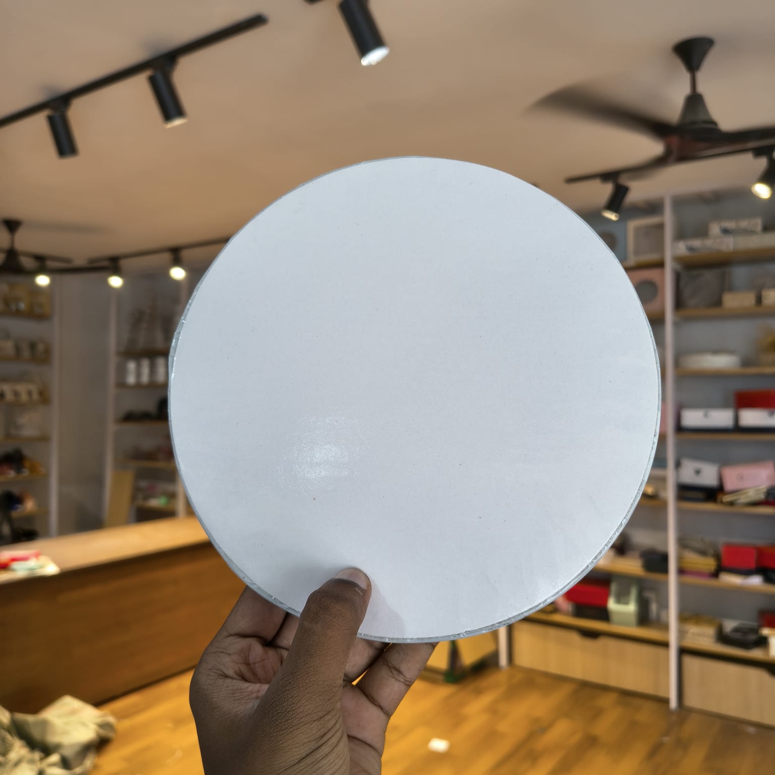 A hand holding a large, plain white circular board or disc in a retail store setting with wooden floors and shelves in the background.